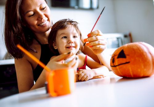A woman and child are painting a small pumpkin with a jack-o'-lantern face, using brushes and enjoying a creative activity.