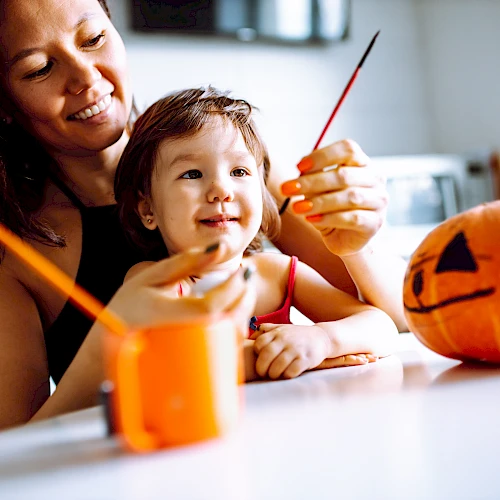 A woman and child are painting a small pumpkin with a jack-o'-lantern face, using brushes and enjoying a creative activity.