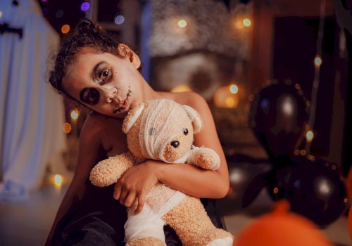 A child with face paint holds a teddy bear, surrounded by Halloween decorations and balloons, creating a festive atmosphere.