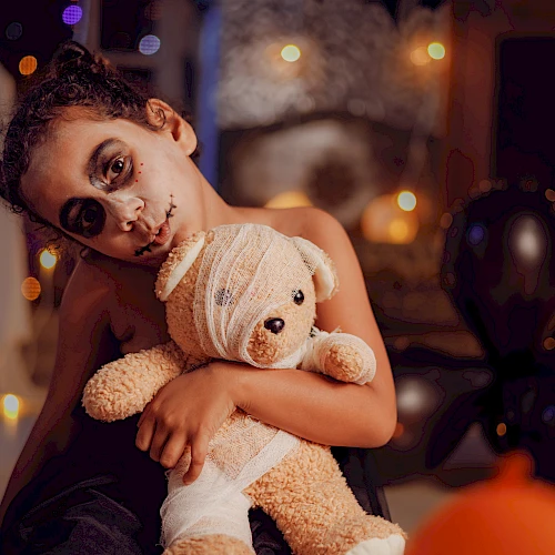 A child with face paint holds a teddy bear, surrounded by Halloween decorations and balloons, creating a festive atmosphere.