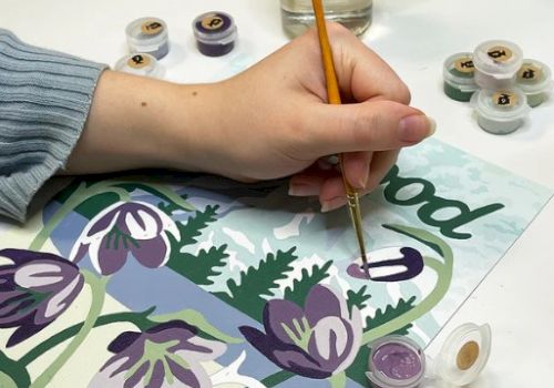 A person paints floral artwork, using brushes and small paint pots to color purple and green flowers on a page.
