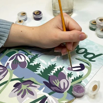 A person paints floral artwork, using brushes and small paint pots to color purple and green flowers on a page.