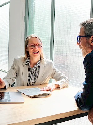 A smiling woman in a light blazer sits at a desk with a laptop, talking to a man in a blue suit across from her in a bright, modern office.