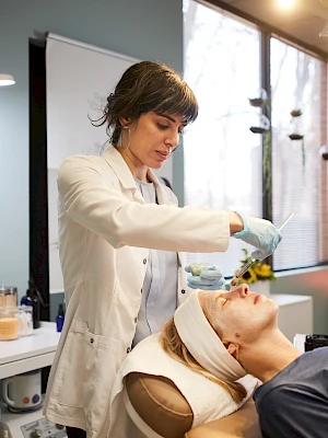A skincare professional applies a facial treatment on a client in a treatment room, with beauty equipment visible around them.