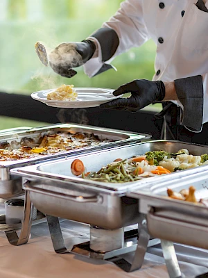 A person in a chef uniform is serving food from buffet trays at a catered event, with various dishes visible on the table.
