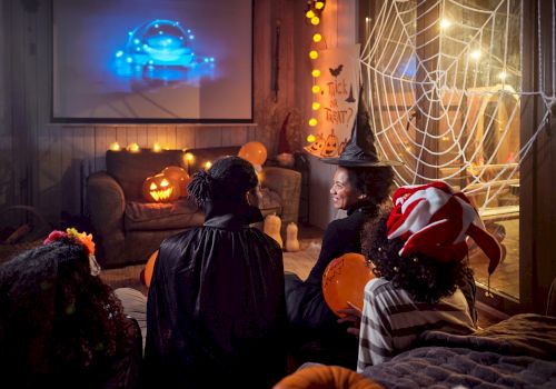 A group in Halloween costumes watches a movie in a decorated room with spider webs and pumpkins.