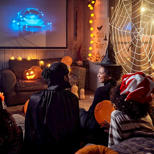 A group in Halloween costumes watches a movie in a decorated room with spider webs and pumpkins.