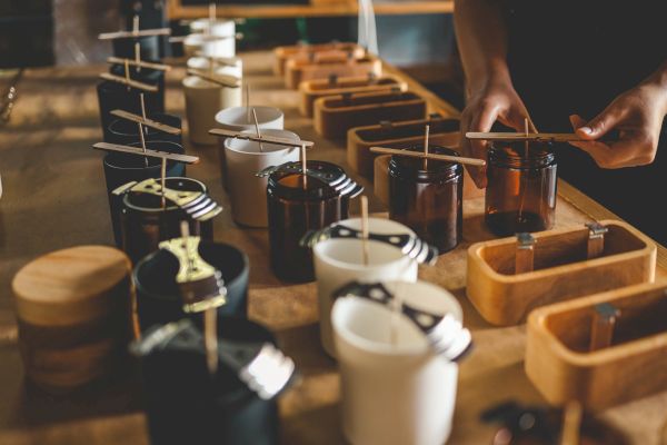 The image shows a person making candles, with several jars and wicks arranged on a table, likely in a candle-making workshop.