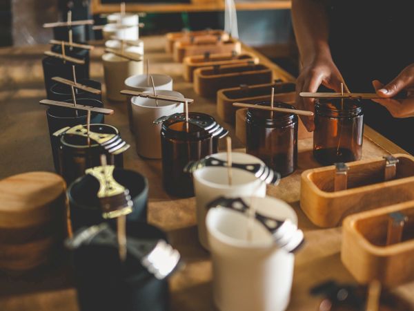 The image shows a person making candles, with several jars and wicks arranged on a table, likely in a candle-making workshop.