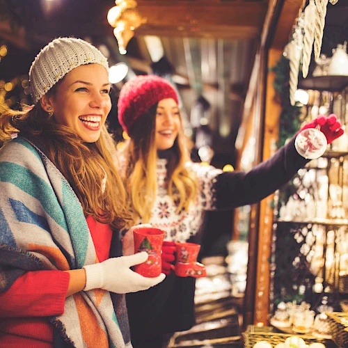 Two women in winter attire, with mugs in hand, enjoying a festive market surrounded by lights and holiday decorations.