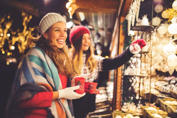 Two women in winter attire enjoy hot drinks and browse festive decorations at an outdoor market stall, surrounded by lights.