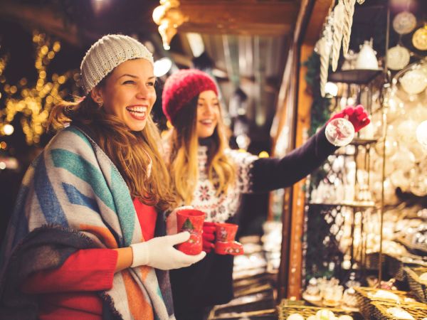 Two women in winter attire enjoy hot drinks and browse festive decorations at an outdoor market stall, surrounded by lights.