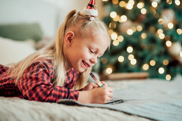 A child in a festive setting writes in a notebook, with a Christmas tree in the background, conveying a cozy holiday atmosphere.