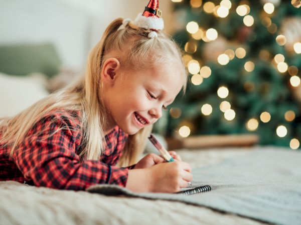 A child in a festive setting writes in a notebook, with a Christmas tree in the background, conveying a cozy holiday atmosphere.