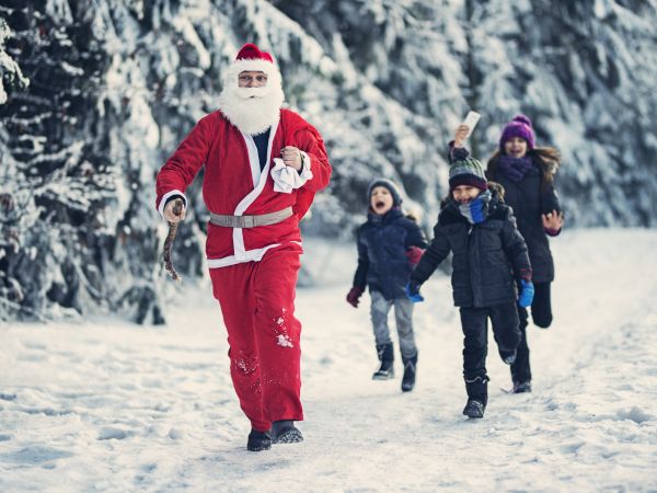 A person in a Santa suit walks in snow, followed by smiling children carrying letters, surrounded by snowy trees.