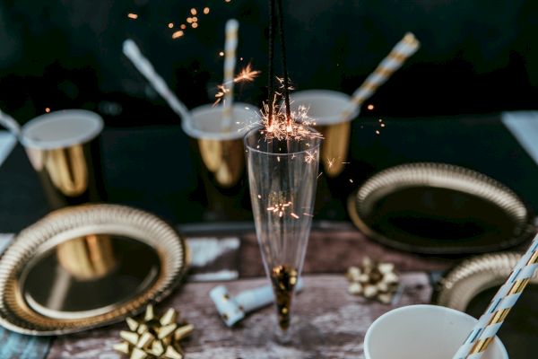 A festive table with gold plates, cups, straws, and a champagne glass with a sparkler lit inside, alongside small golden bows.