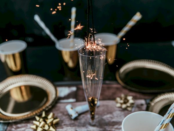 A festive table with gold plates, cups, straws, and a champagne glass with a sparkler lit inside, alongside small golden bows.