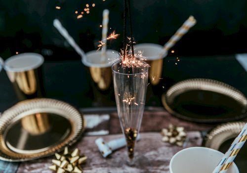 A festive scene with a sparkler in a glass, surrounded by gold-themed plates, cups, and ribbons, creating a celebratory atmosphere.