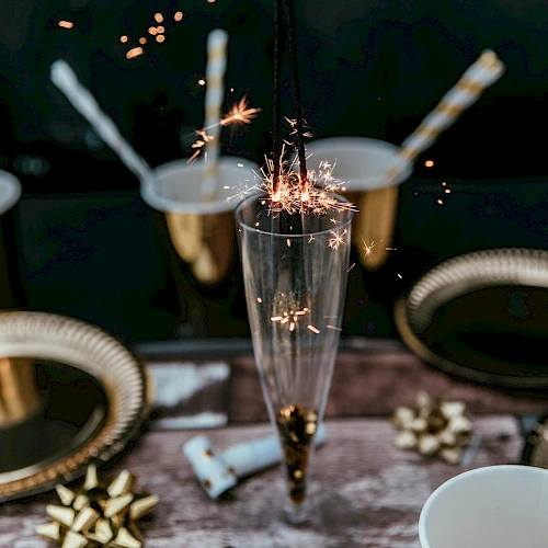 A festive scene with a sparkler in a glass, surrounded by gold-themed plates, cups, and ribbons, creating a celebratory atmosphere.
