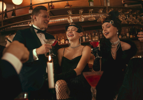 A 1920s-themed party in a bar: women and men in formal attire toast with cocktails and laugh under warm lights.
