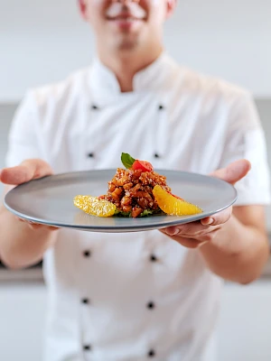 A chef in a white uniform presents a plated dish, featuring a colorful and artistic food presentation on a gray plate.