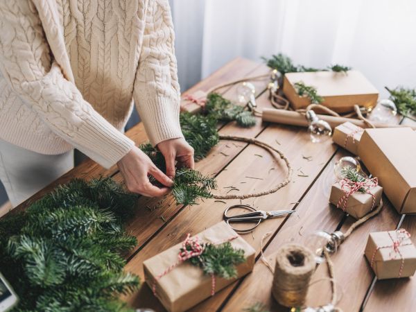 A person is crafting a wreath with greenery on a table, surrounded by wrapped gifts and twine in a cozy setting.
