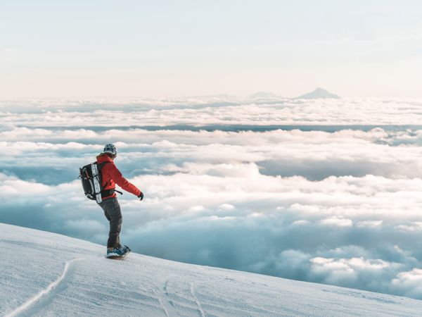 A snowboarder in a red jacket stands on a snowy slope above the clouds, with mountains visible in the distance under a clear sky.