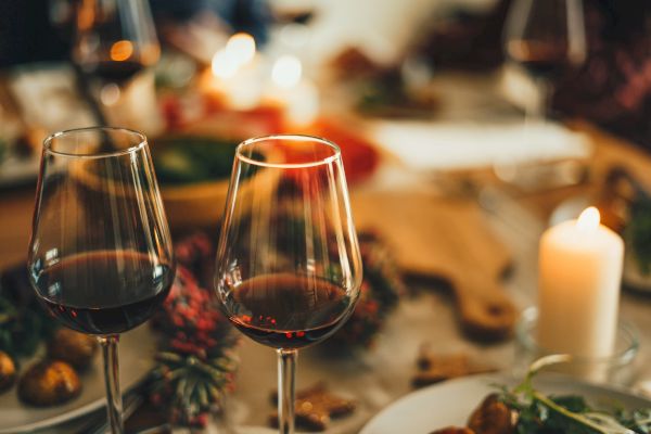 The image shows two glasses of red wine on a dining table with a candle, food, and festive decorations in a warm, cozy ambiance.
