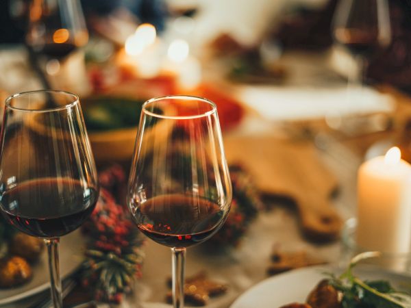 The image shows two glasses of red wine on a dining table with a candle, food, and festive decorations in a warm, cozy ambiance.