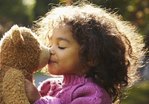 A child in a pink sweater is happily kissing a teddy bear outdoors in the sunlight.