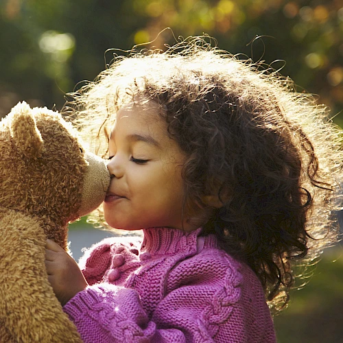 A child in a pink sweater is happily kissing a teddy bear outdoors in the sunlight.
