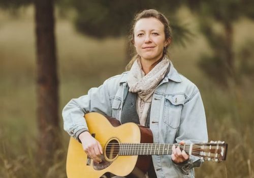 A person in a denim jacket is outdoors, playing an acoustic guitar. The background is a blurred natural setting.