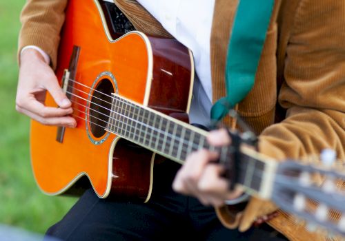 A person playing an acoustic guitar, wearing a brown jacket and white shirt, is featured in the image.