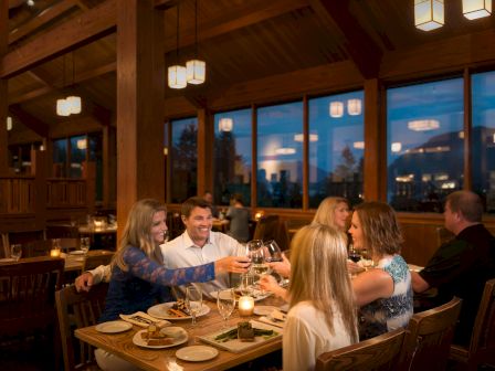 A group of people are dining in a warmly lit restaurant, toasting with glasses, while enjoying a meal at a wooden table.
