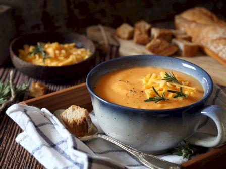 A bowl of creamy soup garnished with cheddar and rosemary, served with bread and croutons on a wooden tray with a striped cloth.