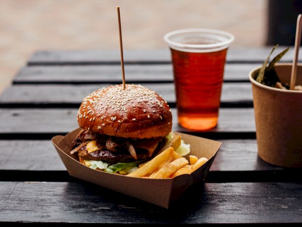 A sesame bun burger with lettuce, cheese, and patty on a tray of fries, a red cup of soda, and a brown takeout container on a wooden table.