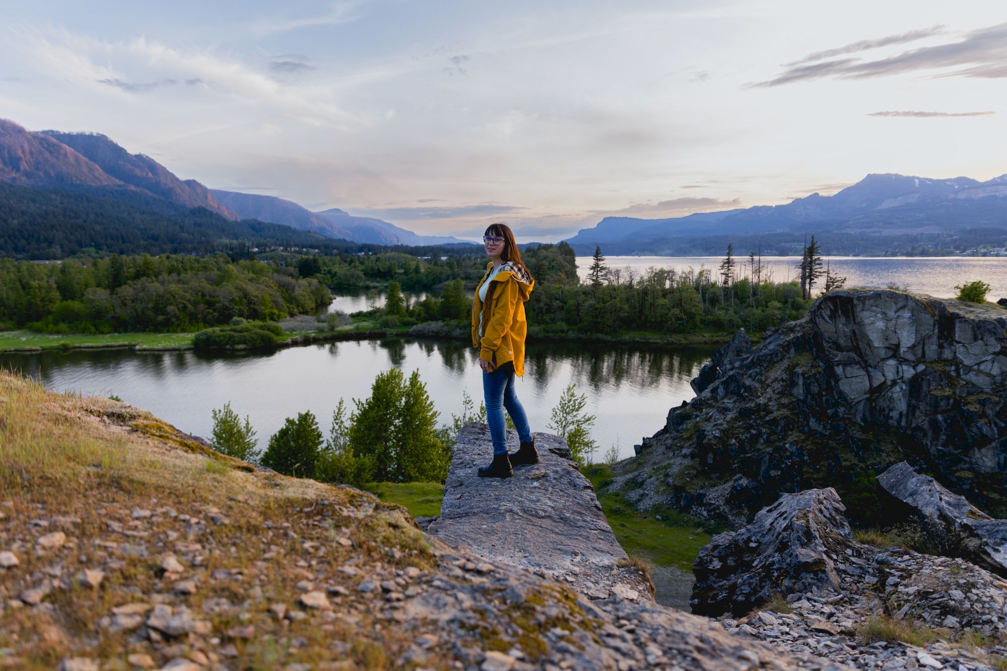 A person in a yellow jacket stands on a rock ledge overlooking a scenic landscape with mountains, trees, and a lake at sunset.