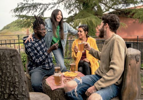 A group of four friends enjoying drinks and snacks outdoors, sitting on wooden stools around a table, with trees in the background.