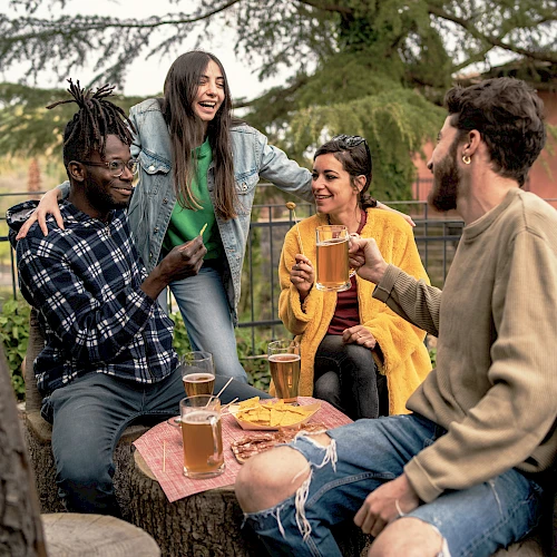 A group of four friends enjoying drinks and snacks outdoors, sitting on wooden stools around a table, with trees in the background.