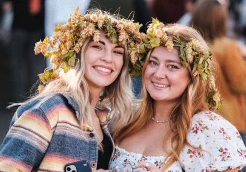 Two people are smiling, wearing flower crowns. They appear to be at an outdoor event, chatting and enjoying themselves in a lively atmosphere.