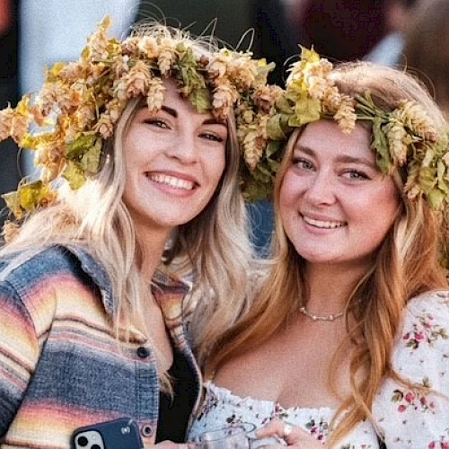 Two people are smiling, wearing flower crowns. They appear to be at an outdoor event, chatting and enjoying themselves in a lively atmosphere.