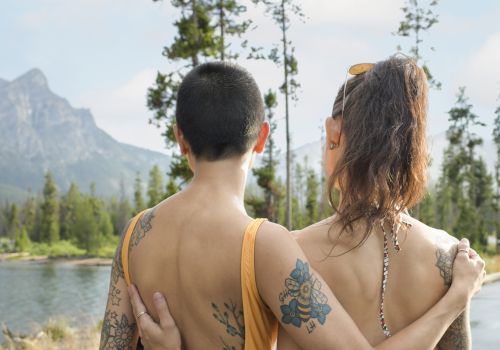 Two people with tattoos are embracing, facing a scenic view of mountains and a lake.