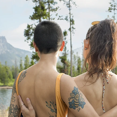 Two people with tattoos are embracing, facing a scenic view of mountains and a lake.