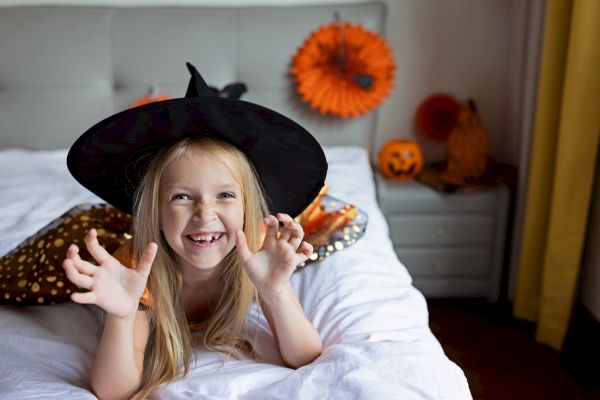 A smiling child wearing a witch hat poses playfully on a bed with Halloween decorations in the background, including pumpkins.