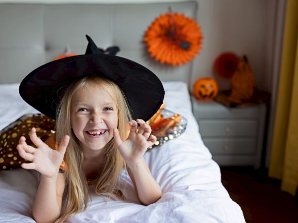 A smiling child wearing a witch hat poses playfully on a bed with Halloween decorations in the background, including pumpkins.