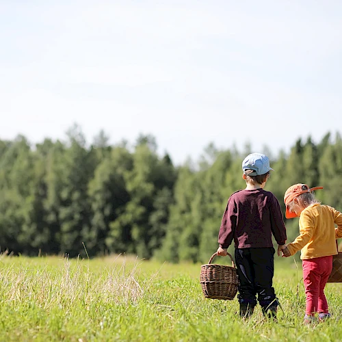 Two children in a grassy field, holding baskets and wearing hats, with trees and hay bales in the background.