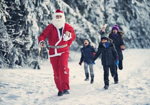 A person in a Santa suit is playfully running in the snow, followed by excited children dressed in winter clothing.