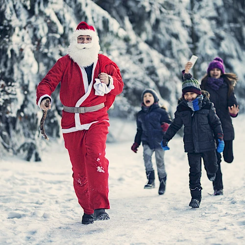 A person in a Santa suit is playfully running in the snow, followed by excited children dressed in winter clothing.