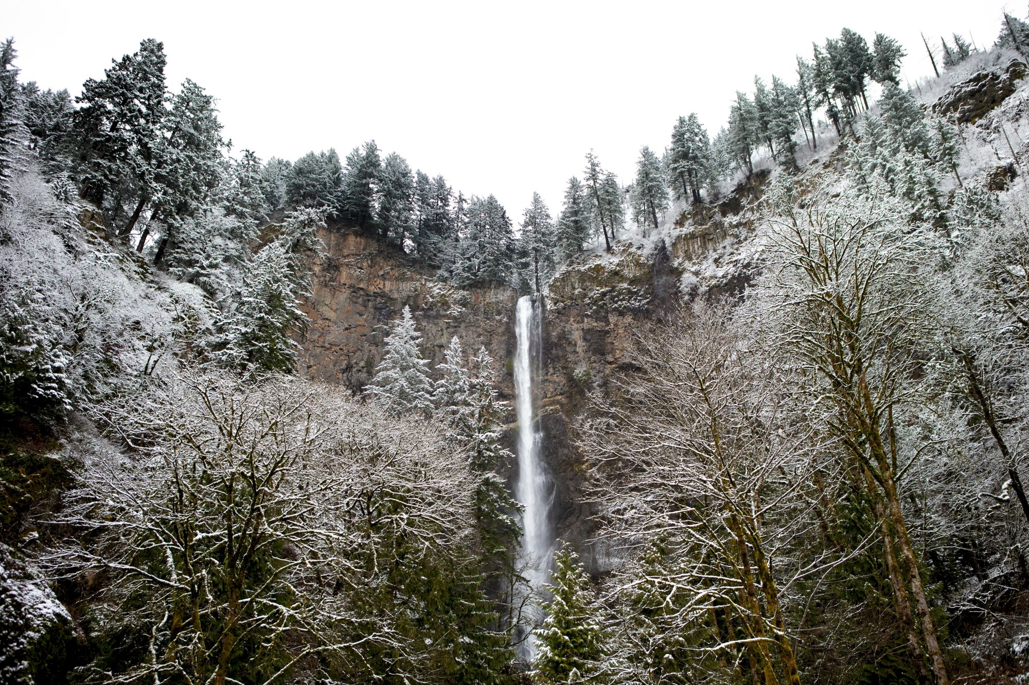 A tall waterfall cascades down a cliff surrounded by snowy trees and a forested landscape under a cloudy sky.