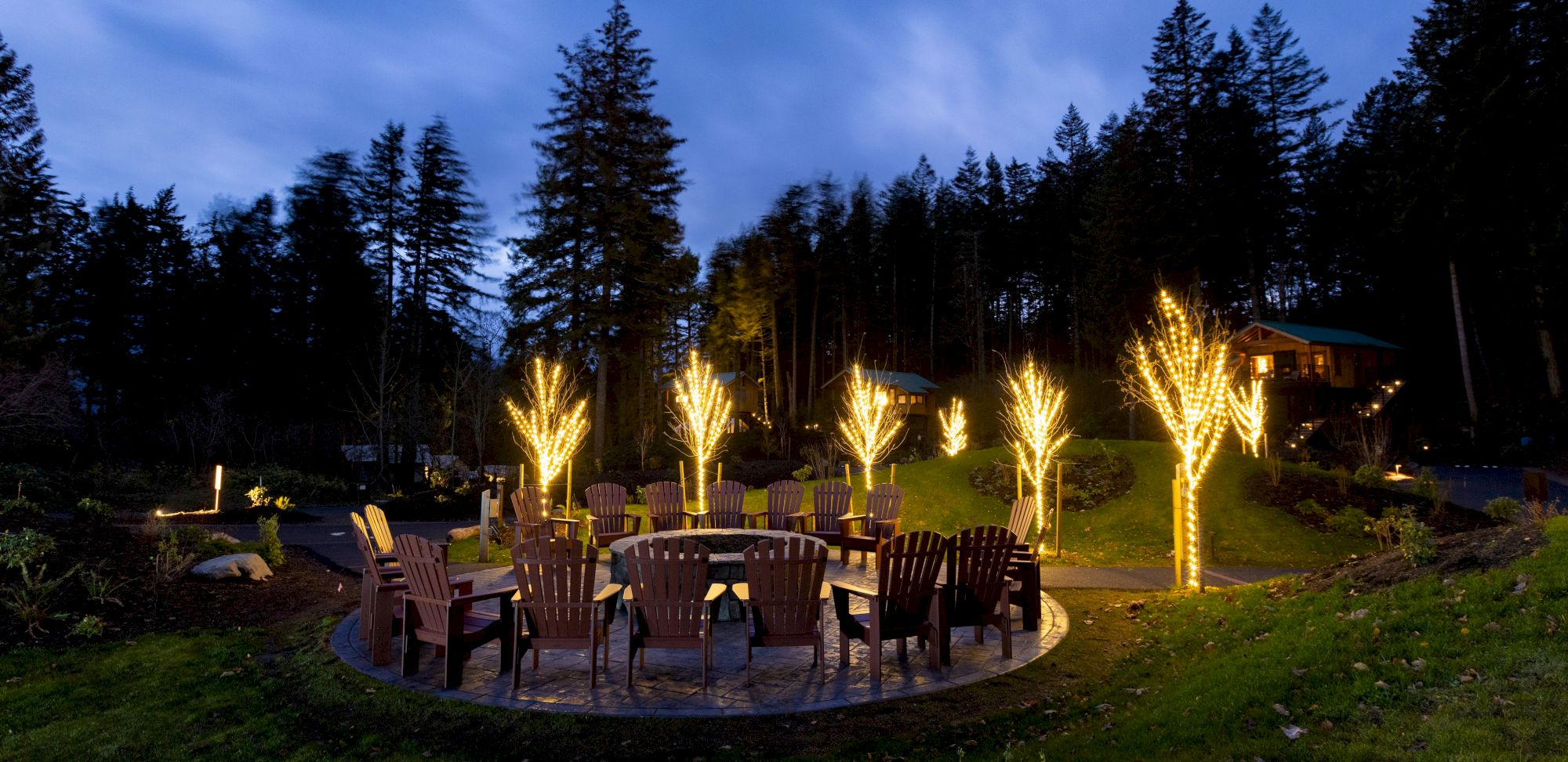 A circular outdoor seating area with wooden chairs surrounds trees wrapped in glowing lights at dusk, set against a forest backdrop.